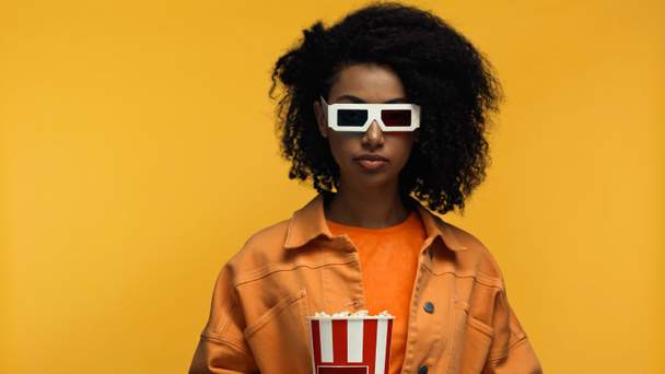 młoda Afroamerykanka w okularach 3d trzymając popcorn wiadro izolowane na żółto - Zdjęcie, obraz