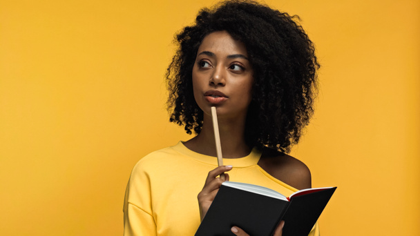 zamyšlená afro-americká žena drží pero a notebook izolované na žluté  - Fotografie, Obrázek