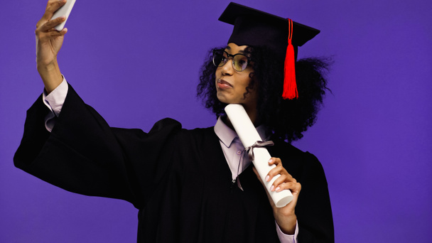 joven africano americano estudiante en gafas y graduación gorra tomando selfie aislado en púrpura  - Foto, Imagen