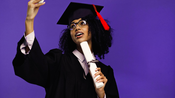 boldog afro-amerikai diák fogszabályzóval ballagási sapkában és köntösben szelfit készít hengerelt diplomával elszigetelt lila  - Fotó, kép