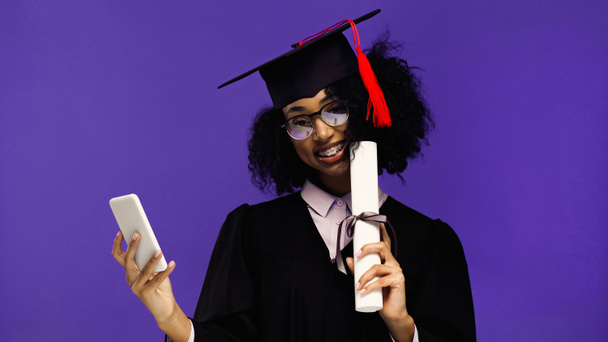 усміхнений афроамериканський студент з брекетами у випускній шапці та сукні, що тримає смартфон та диплом ізольовані на фіолетовому
  - Фото, зображення