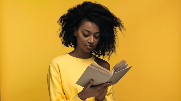 curly african american woman reading book isolated on yellow - Фото, изображение