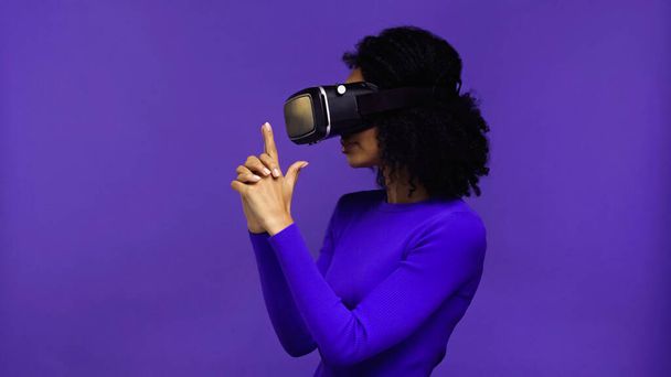 curly african american woman in vr headset showing finger gun gesture with hands isolated on purple - Fotó, kép