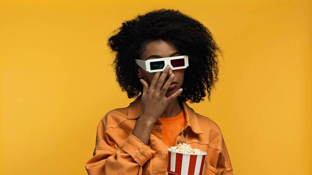 sad young african american woman in 3d glasses holding popcorn bucket while rubbing eye isolated on yellow - Fotó, kép