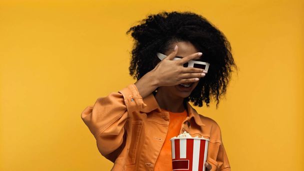 assustado jovem afro-americano mulher em aparelho e 3d óculos segurando pipoca balde isolado no amarelo - Foto, Imagem
