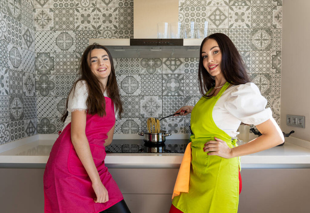 Mujeres jóvenes y adultas felices en delantales sonriendo y mirando a la cámara mientras hierve espaguetis en la cocina moderna en casa - Foto, imagen