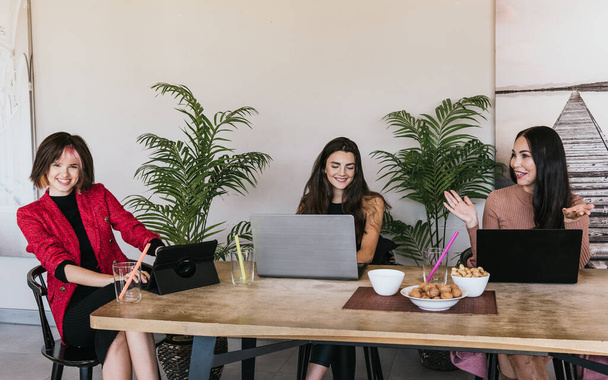 Gleeful donne alla moda in abiti casual utilizzando dispositivi digitali e sorridente mentre seduto al tavolo della caffetteria e parlando durante il lavoro remoto - Foto, immagini