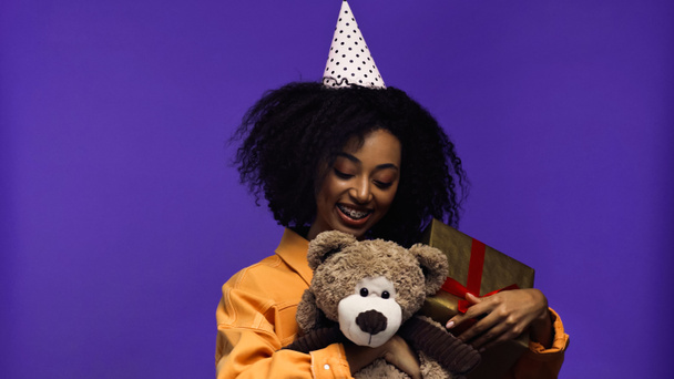 veselý africký Američan žena s rovnátka a strana čepice držení zabalené dárkové krabice a medvídek izolované na fialové - Fotografie, Obrázek