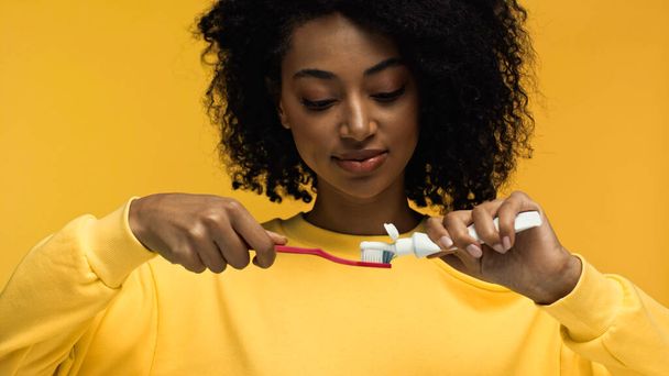 mulher afro-americana espremendo pasta de dentes em escova de dentes isolada em amarelo - Foto, Imagem
