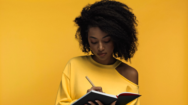 lockige Afroamerikanerin schreibt in Notizbuch isoliert auf gelb  - Foto, Bild