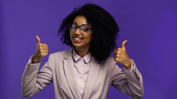 szczęśliwy afrykański amerykański bizneswoman w okulary i marynarka pokazując kciuki w górę odizolowany na fioletowy  - Zdjęcie, obraz