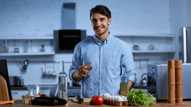 happy young man holding glass of white wine near table with fresh ingredients - Fotó, kép