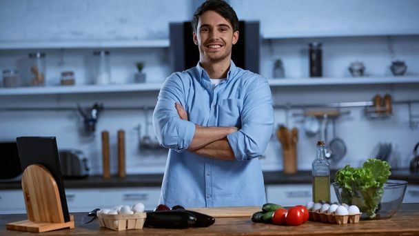 sonriente hombre mirando a la cámara mientras está de pie con los brazos cruzados cerca de la mesa con ingredientes frescos - Foto, imagen