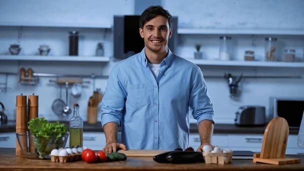 sonriente hombre mirando a la cámara mientras está de pie cerca de la mesa con ingredientes frescos
 - Foto, imagen