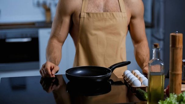 cropped view of shirtless man in apron standing near frying pan and chicken eggs - Фото, изображение