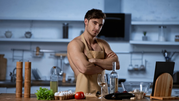 bez koszulki mężczyzna w fartuchu pozowanie z skrzyżowanymi ramionami patrząc na aparat fotograficzny w kuchni  - Zdjęcie, obraz