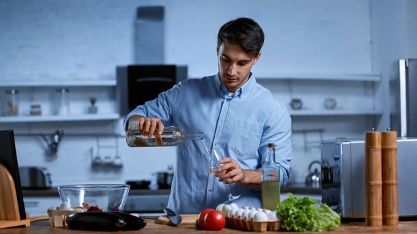young man pouring white wine in glass near table with fresh vegetables - Foto, afbeelding