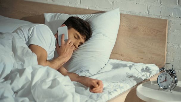 hombre con los ojos cerrados hablando en el teléfono móvil en la cama cerca del despertador en la mesita de noche - Foto, imagen