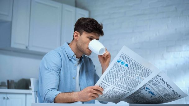 homem de camisa azul bebendo café e lendo jornal da manhã na cozinha - Foto, Imagem