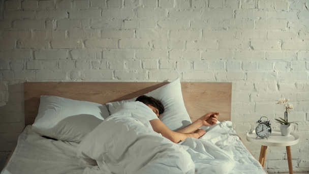 man sleeping under white blanket near vintage alarm clock and orchid on bedside table - Zdjęcie, obraz
