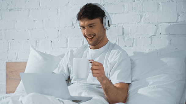 cheerful man in headphones holding cup near laptop in bed - Photo, Image