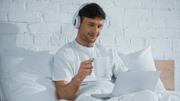 sorrindo homem em fones de ouvido segurando copo ao usar laptop na cama - Foto, Imagem
