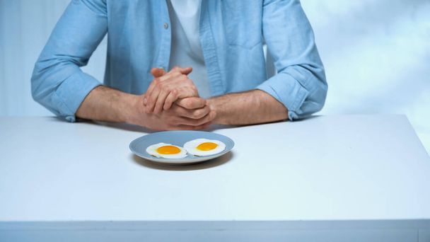 cropped view of man sitting with clenched hands near plate with fried eggs - Fotografie, Obrázek