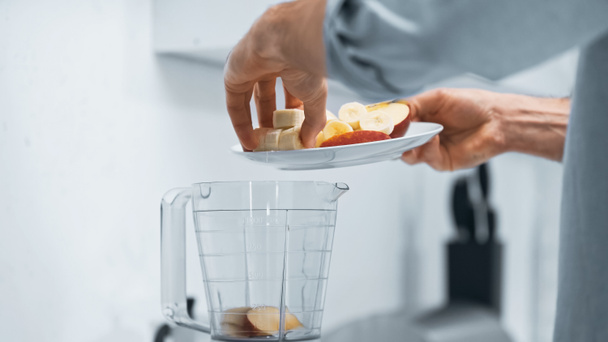 cropped view of man adding cut apple and banana into bowl of blender - Fotografie, Obrázek