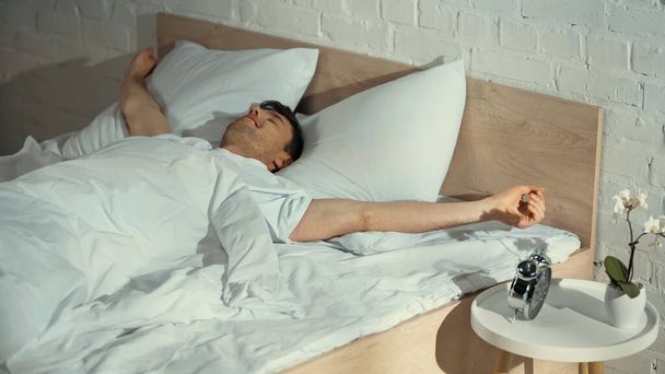 проснувшийся мужчина улыбается, растягиваясь в постели возле будильника на тумбочке - Фото, изображение