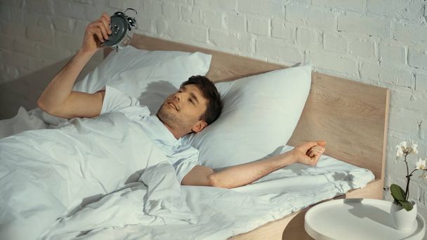 sorrindo homem olhando para relógio de alarme vintage enquanto deitado na cama de manhã - Foto, Imagem