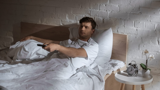 目覚まし時計の近くのベッドでテレビを見ながらチャンネルをクリックする男とベッドサイドテーブルで蘭 - 写真・画像