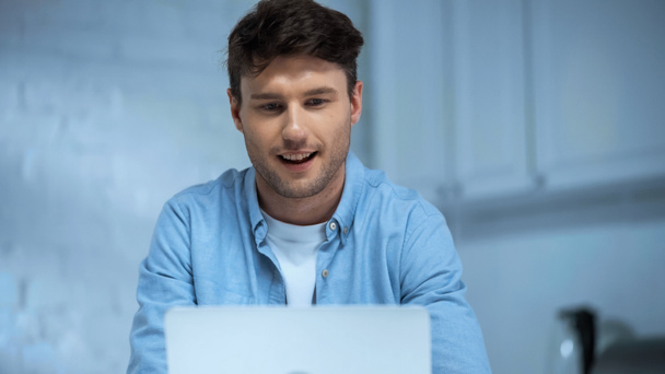 smiling man in blue shirt working on laptop in kitchen - 写真・画像