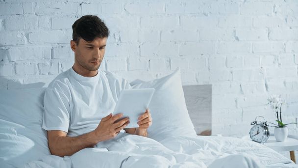 hombre sentado en la cama cerca de reloj despertador vintage y el uso de tableta digital - Foto, imagen