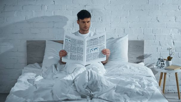 мужчина читает газету, сидя на кровати под белым одеялом - Фото, изображение