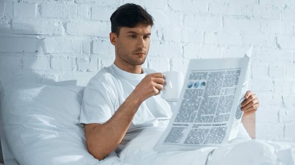 man with cup reading morning newspaper while sitting on bed in morning - Foto, imagen