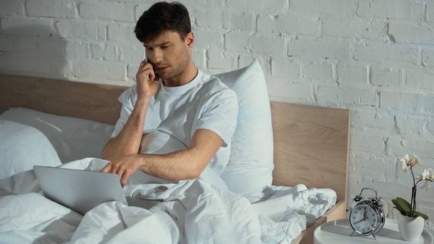 man pointing at laptop while talking on smartphone in bed - Photo, Image