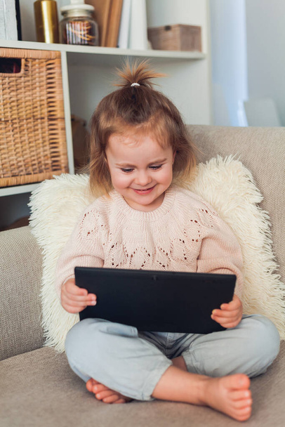 Koulu verkossa. Suloinen pieni tyttö 3-vuotias istuu sohvalla viihtyisässä talossa ja puhuu videopuhelulla. Muotokuva tunteikkaasta lapsesta, jolla on tabletti käsissään. Kotiin. - Valokuva, kuva