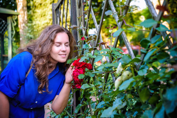 Beautiful girl smells the roses. Woman doing garden work sniffing at the roses at beautifully sunny day. - Photo, Image