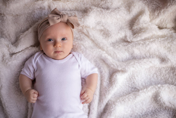 Baby-Mädchen auf einer weißen Decke in weißer Kleidung auf dem Bauch liegend und lächelnd, in die Kamera blickend, Baby-Morgen, Baby-Zeug-Konzept. Platz für Text. - Foto, Bild