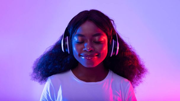 ワイヤレスヘッドフォンで音楽を聴く魅力的な若い黒人女性,ネオンライトで閉じた目で笑顔,パノラマ - 写真・画像