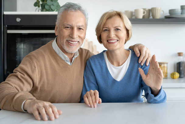 Portret uśmiechniętej pary seniorów robiącej zdjęcia i patrzących w kamerę siedzącej przy stole w nowoczesnej kuchni. Szczęśliwa rodzina. Komunikacja na odległość z przyjaciółmi i krewnymi - Zdjęcie, obraz