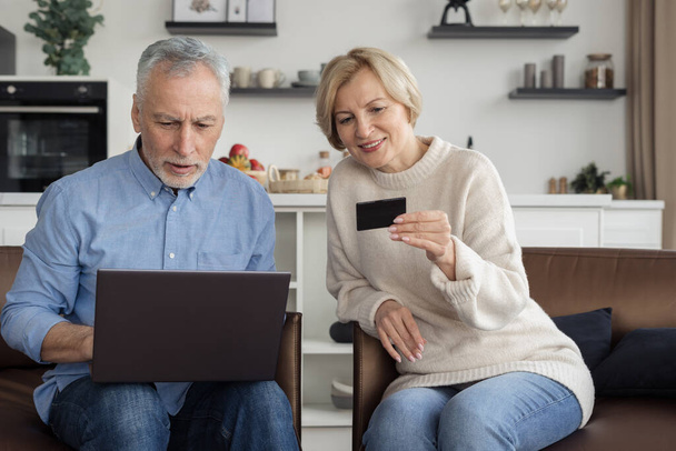 Szczęśliwa para emerytów dokonujących zakupów online za pomocą karty kredytowej i laptopa w nowoczesnym stylu kuchni. Koncepcja zakupów online. Pomysł na konsumpcję - Zdjęcie, obraz
