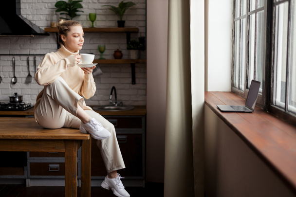 Gelukkige jonge vrouw is in een gezellige huiselijke sfeer, zit aan de keukentafel en houdt een kopje koffie in haar handen - Foto, afbeelding