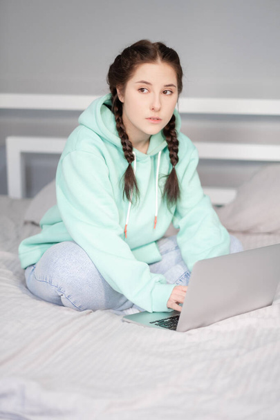 молода брюнетка з двома кісками в бірюзовому светрі і джинсах сидить на ліжку з білими простирадлами з ноутбуком. Працювати або вчитися вдома. карантинні обмеження. віддалена робота, дистанційне навчання
 - Фото, зображення
