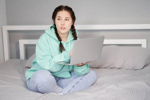 mladá brunetka se dvěma copánky v tyrkysové mikině a džínami sedící na posteli s bílými prostěradly s laptopem. pracovat nebo studovat z domova. karanténní omezení. vzdálená práce, dálkové studium - Fotografie, Obrázek