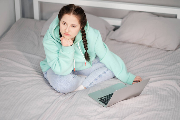 Młoda brunetka z dwoma warkoczami w turkusowej bluzie i dżinsami siedząca na łóżku z białymi prześcieradłami z laptopem. praca lub nauka w domu. ograniczenia kwarantanny. praca na odległość, nauka zdalna - Zdjęcie, obraz