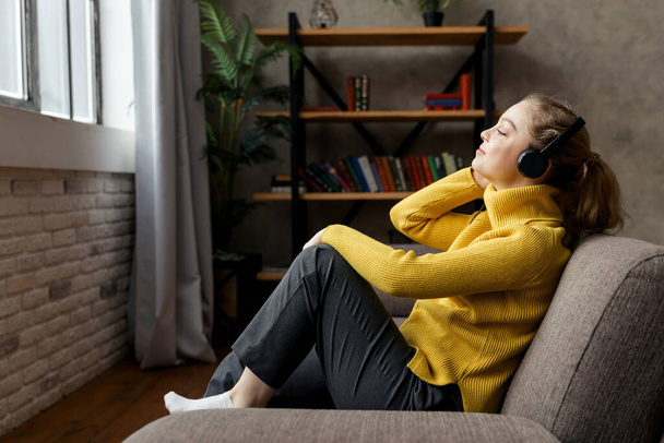 Beautiful pleased woman listening to music in headphones while sitting on sofa at home - Foto, imagen