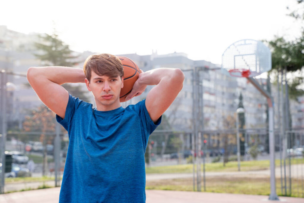 Porträt eines jungen Mannes auf einem Basketballplatz, der einen Basketball hinter seinem Kopf hält. - Foto, Bild