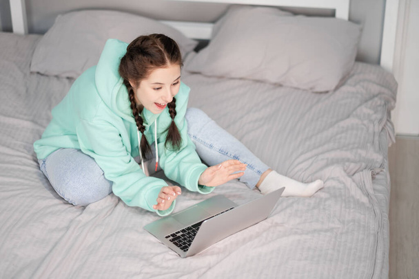 young brunette woman with two pigtails in turquoise hoodie and jeans sitting on a bed with white sheets with laptop. working or studying from home. quarantine restrictions. distant work, remote study - Fotoğraf, Görsel