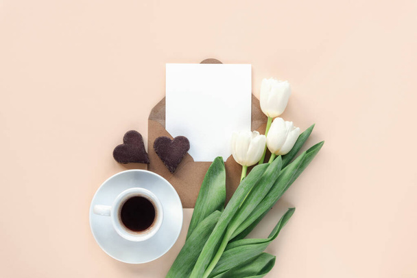 Fehér tulipán, boríték üres lappal, kávéscsésze és szív alakú csokoládé sütik semleges bézs háttérrel. Valentin-nap, anyák napi ünnepi fogalom. Felülnézet, lapos fekvés, mockup. - Fotó, kép